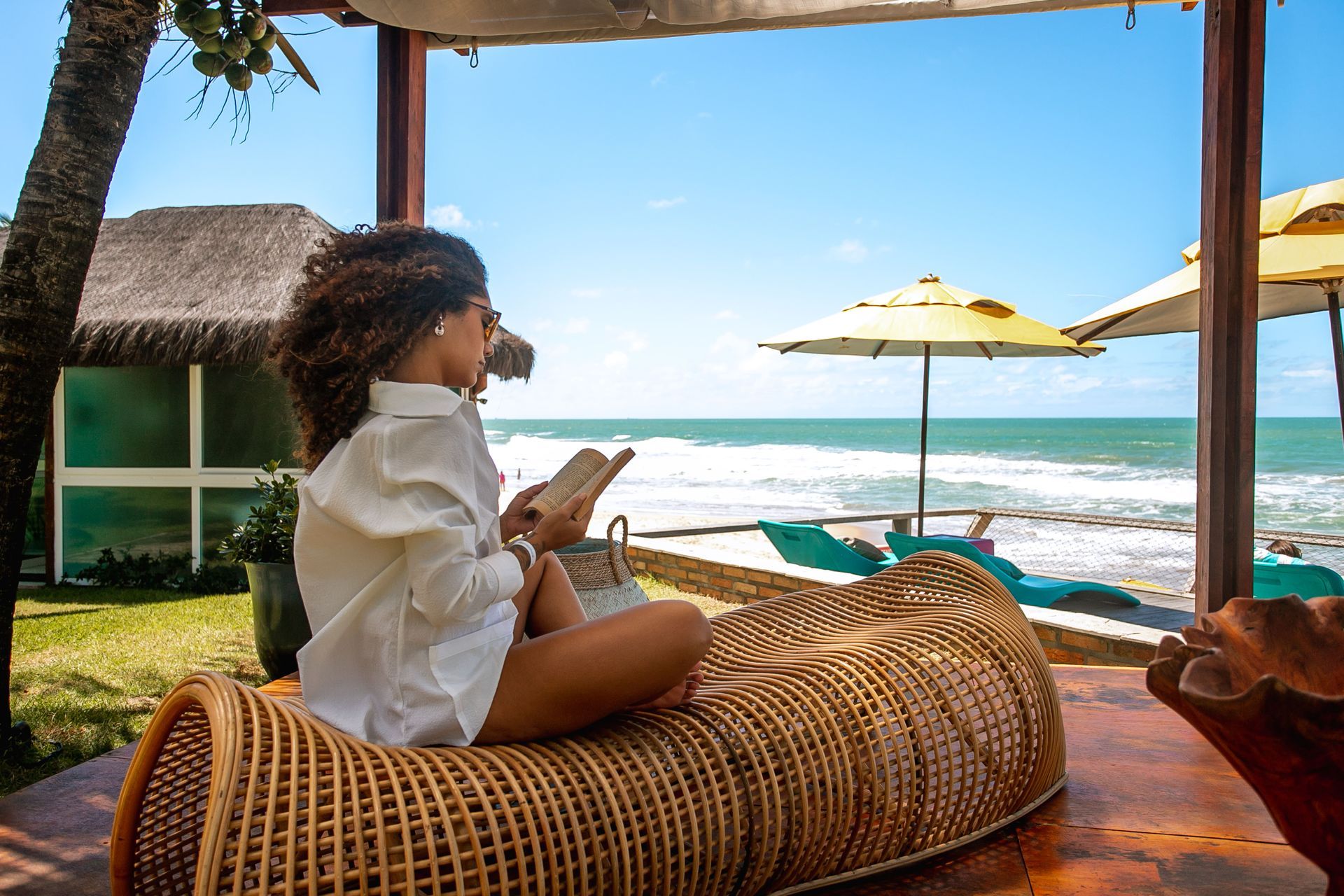 Mulher sentada em espreguiçadeira de fibra natural lendo um livro em frente ao mar, com guarda-sóis amarelos e espreguiçadeiras à beira da praia em um dia ensolarado.