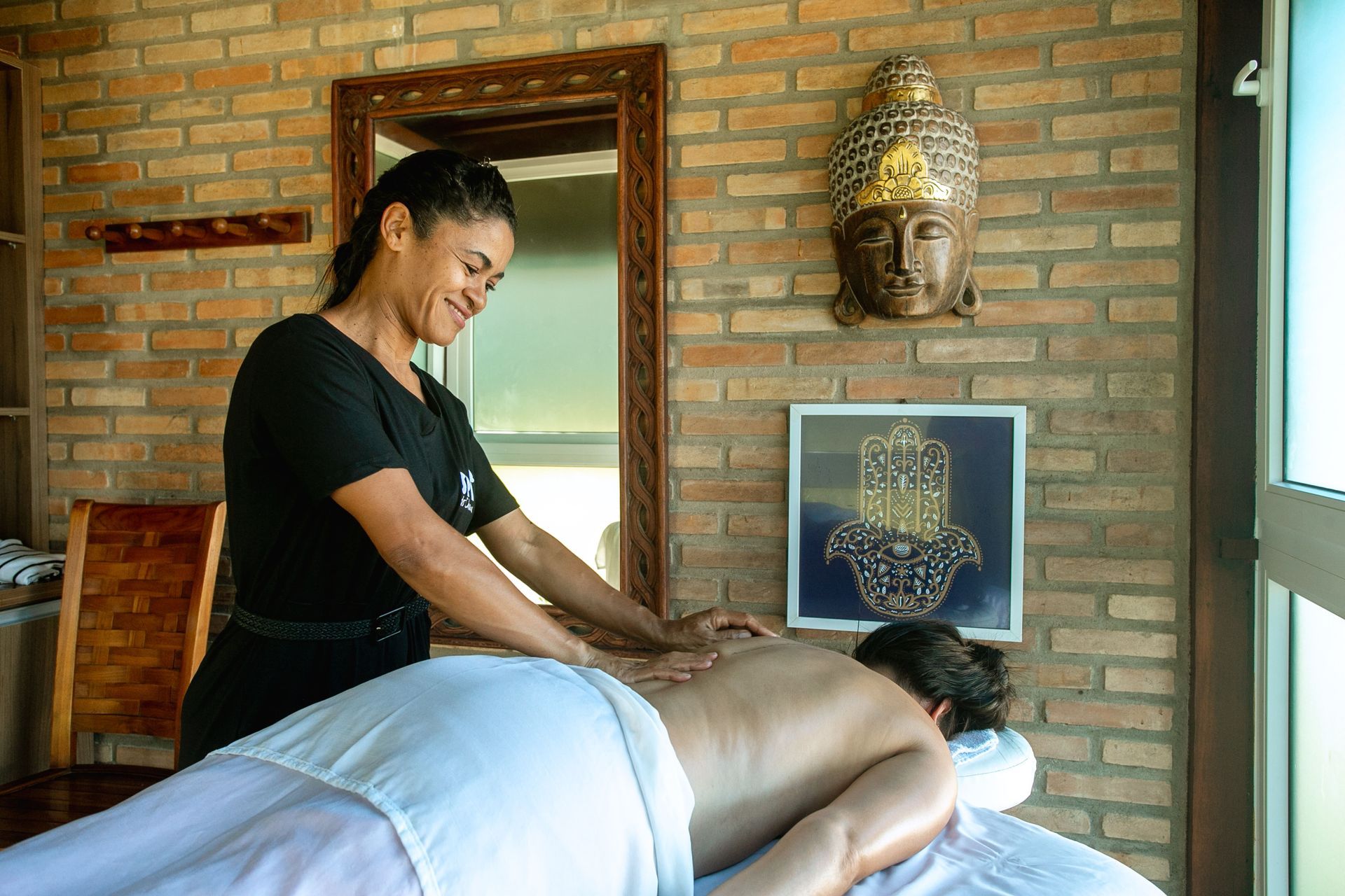 Massagista sorridente realizando massagem relaxante em um spa com decoração zen, parede de tijolos, escultura de Buda e símbolo da mão de Hamsá.