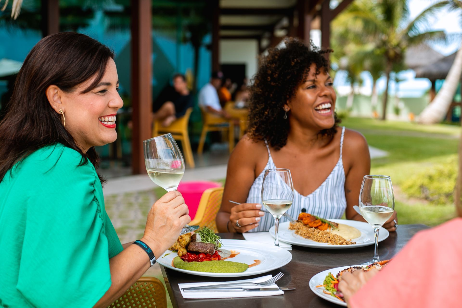 Três mulheres sorrindo e apreciando uma refeição ao ar livre no restaurante do Kembali, com pratos de comida gourmet e taças de vinho. Ao fundo, há mais pessoas sentadas em mesas, e o ambiente é cercado por vegetação tropical.