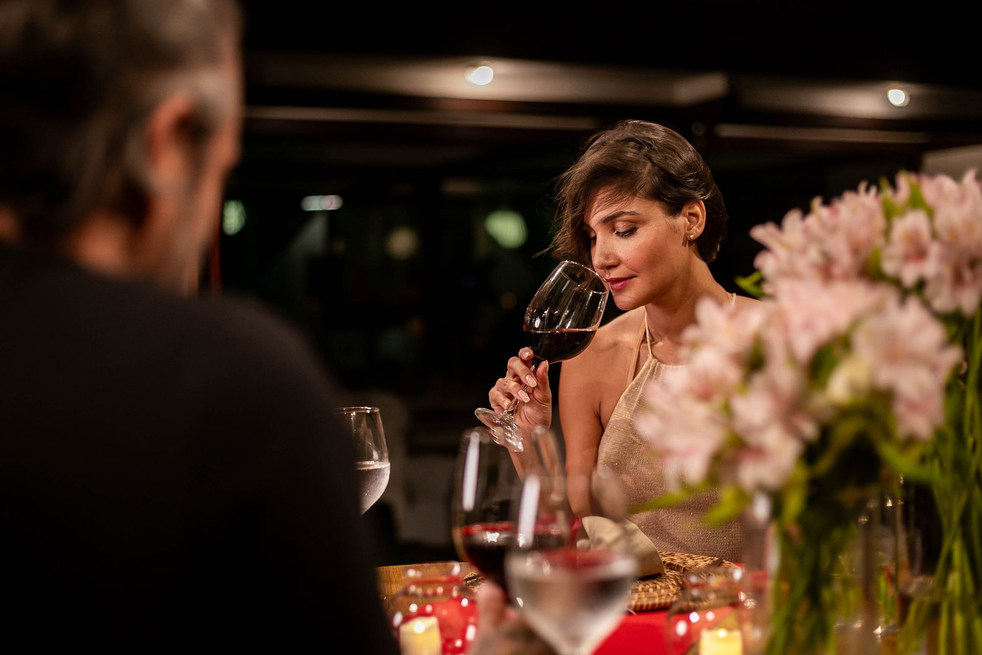 Mulher apreciando uma taça de vinho tinto em um jantar romântico à luz de velas, com flores ao fundo, criando uma atmosfera elegante e intimista.