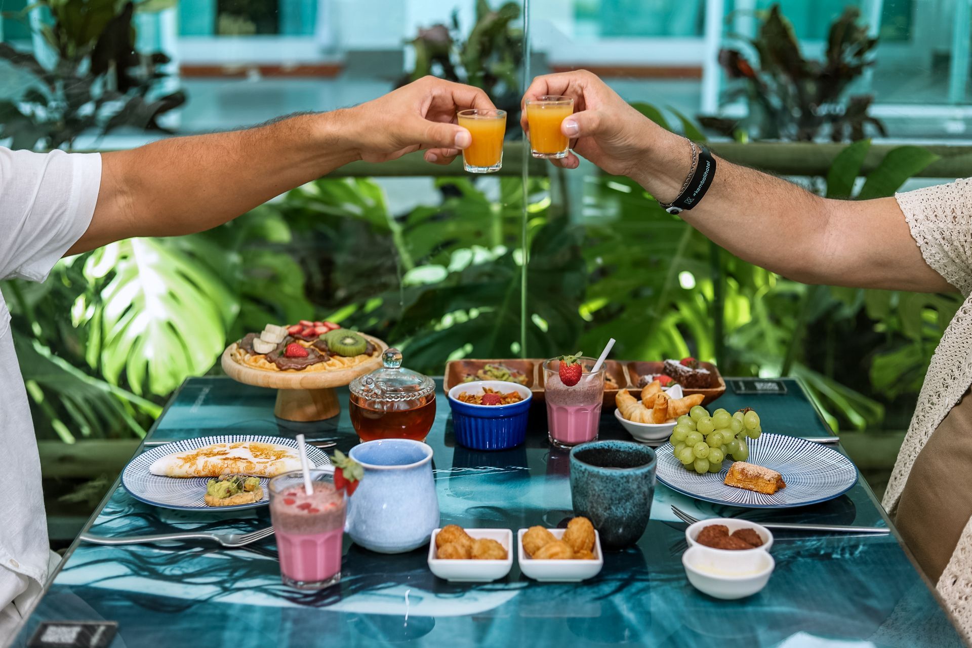 Mesa no restaurante do hotel, com várias opções de pratos do café da manhã, como tapioca, vitamina, mini churros, frutas, waffle e café. Duas pessoas sentada estão brindando com shots de imunidade. Ao fundo, folhas de costela de adão desfocadas.