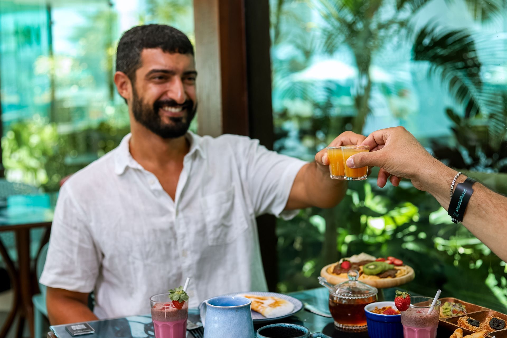 Homem sentado com uma mesa de café da manhã bem servida. Ele sorri e segura um pequeno como de shot amarelo, brindando com outra mão de pessoa que não aparece na foto.
