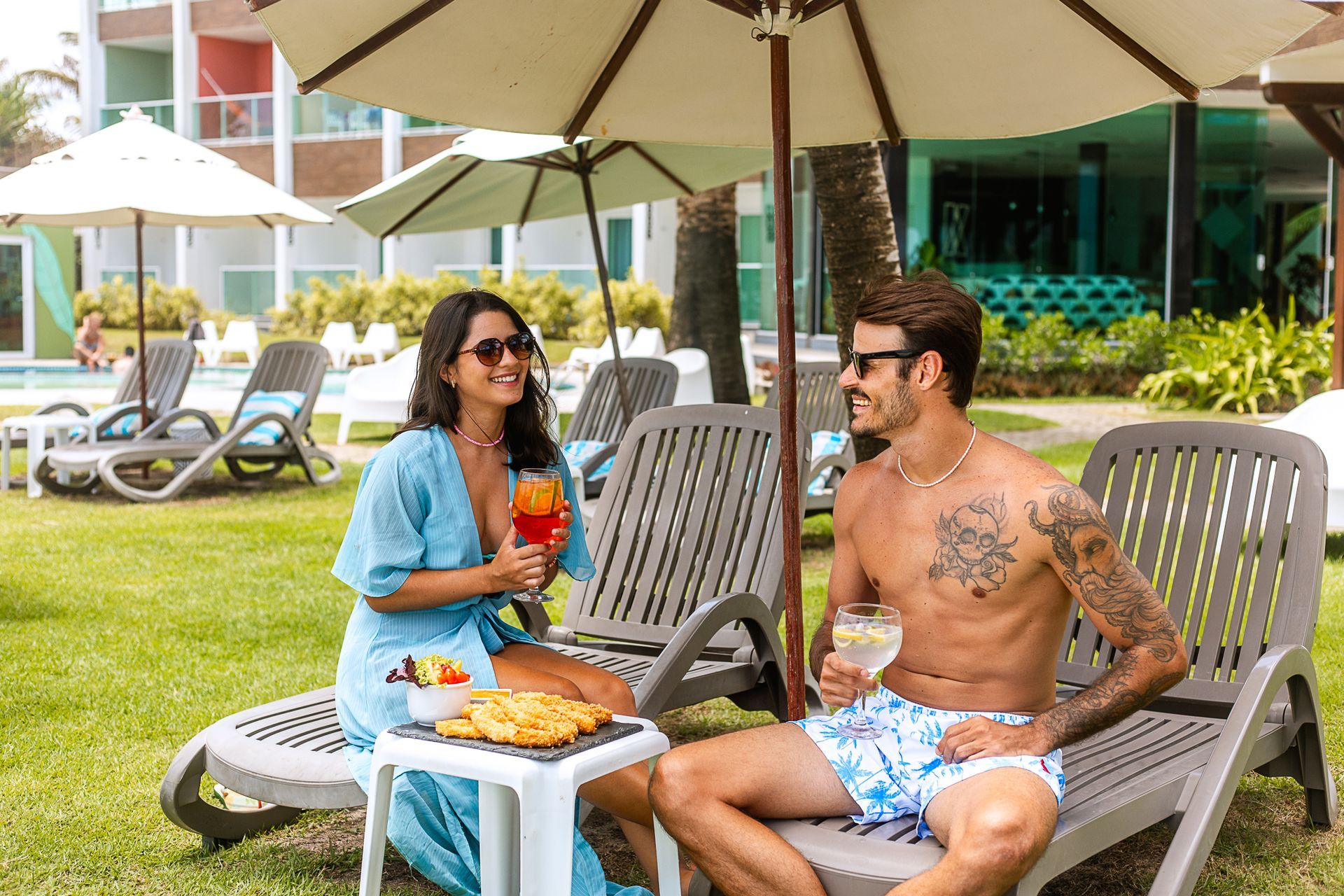 Casal em espreguiçadeiras na área externa; comida, bebida e hotel colorido de fundo.