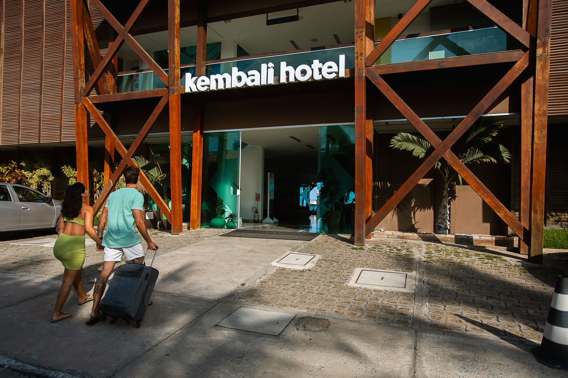 Um casal caminha em direção à entrada do hotel Kembali Hotel. Um homem puxa uma mala preta de rodinhas, enquanto uma mulher o acompanha. A fachada do hotel é moderna, com estruturas de madeira escura e grandes portas de vidro. 