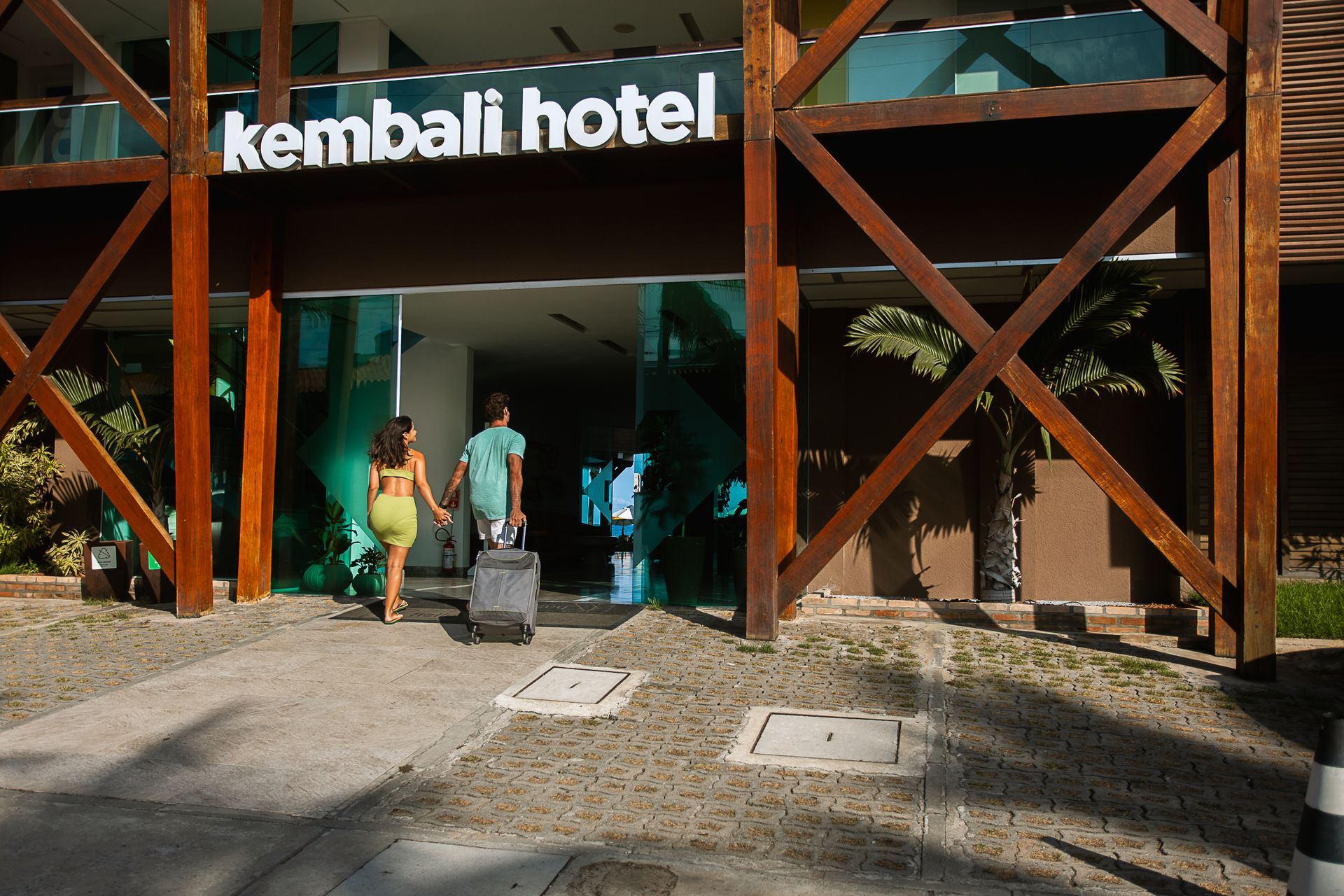 Entrada do Kembali Hotel; casal com mala chegando pela porta de madeira e vidro.