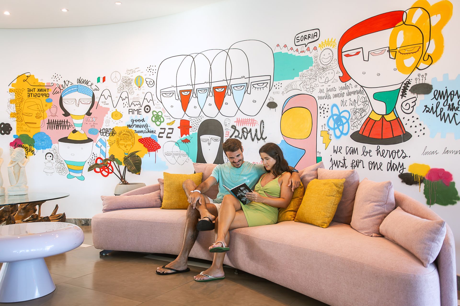 Lounge com sofá rosa; casal sentado em frente a parede com arte colorida do Art Hub.