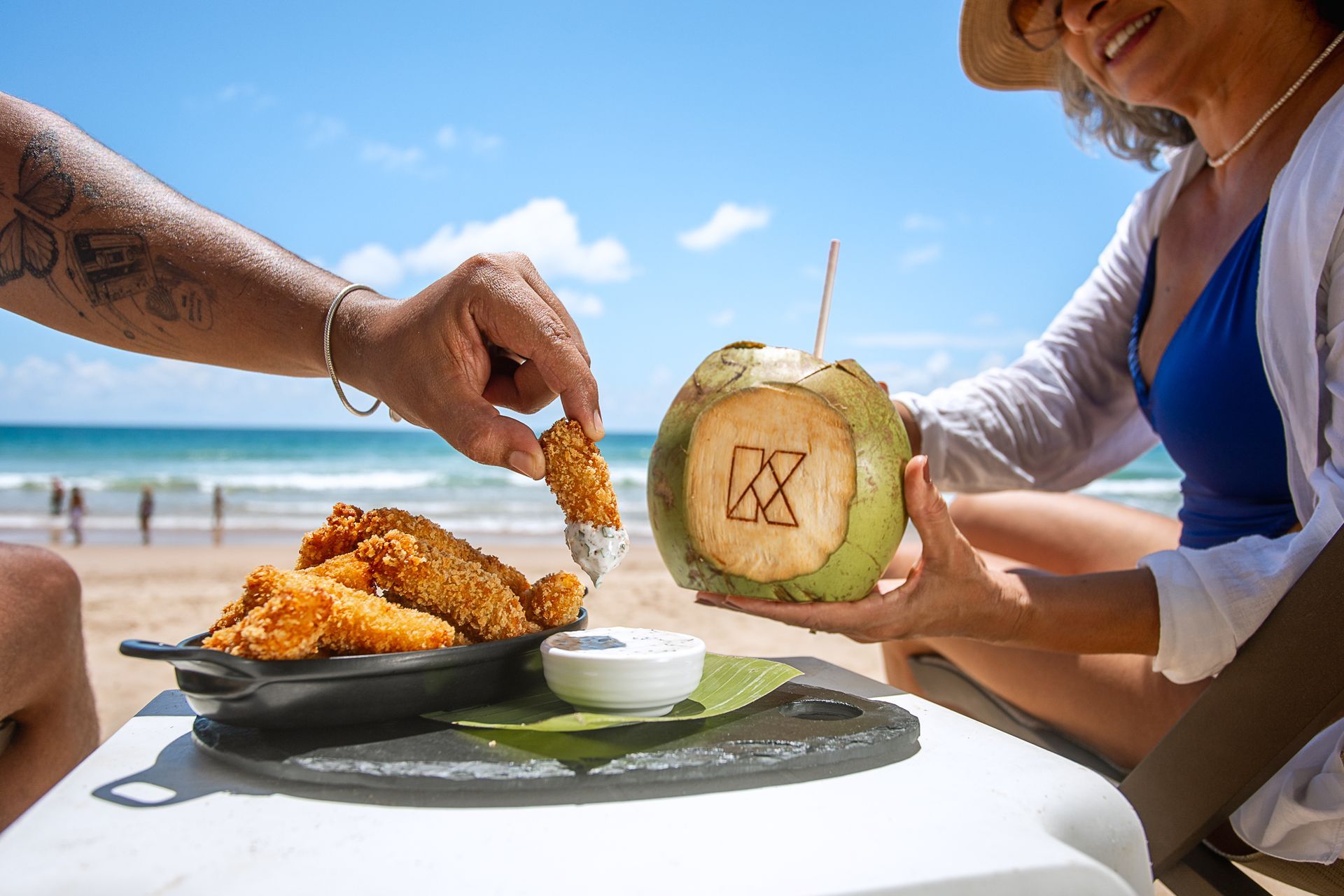 Casal na praia com petiscos à mesa, mulher segurando um coco e o mar ao fundo.