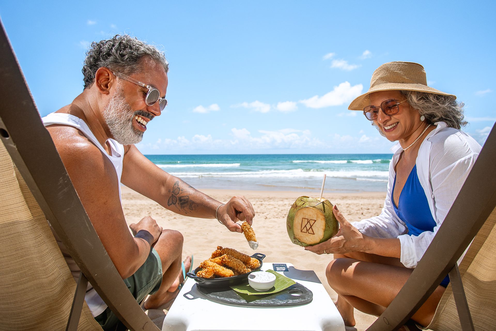 Um casal relaxa em espreguiçadeiras na praia, sob um céu azul. Eles sorriem enquanto compartilham petiscos sobre uma pequena mesa; um deles segura um coco com canudo e o outro pega comida de um prato. O mar e as ondas aparecem ao fundo, criando um clima tranquilo e ensolarado.