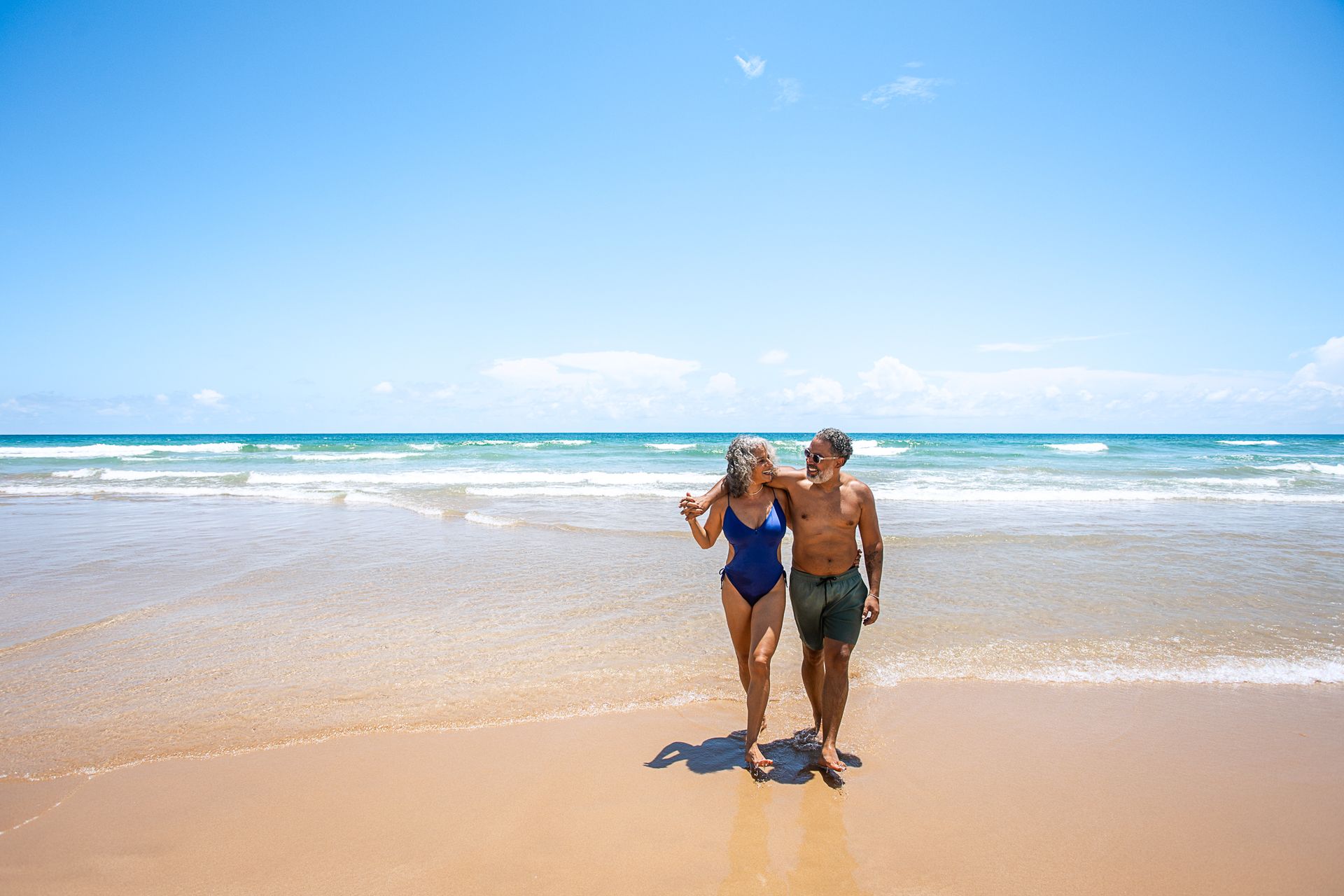 Casal caminhando sorridente na beira do mar em uma praia ensolarada, com o oceano e o céu azul ao fundo.