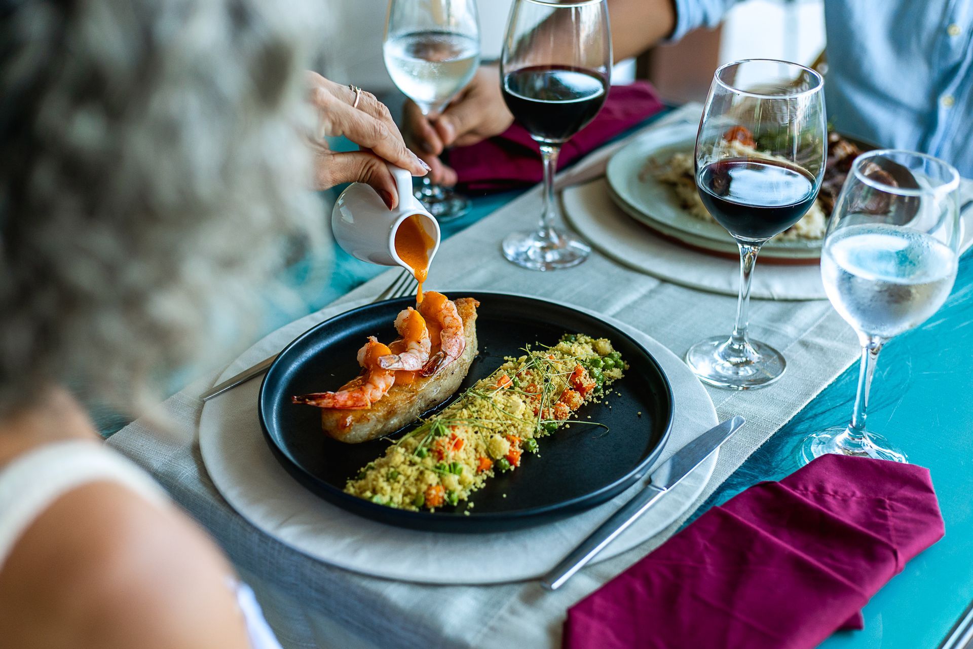 Prato fino: camarão com molho sendo servido, acompanhamento verde, taças de vinho & louça sofisticada