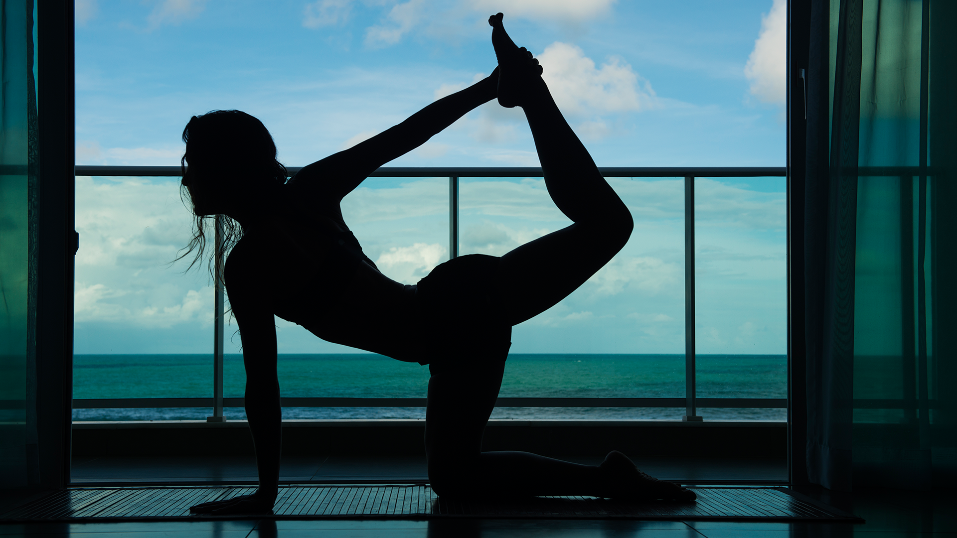 Mulher praticando yoga na varanda do quarto do hotel. Ao fundo, o céu e o mar estão azuis.