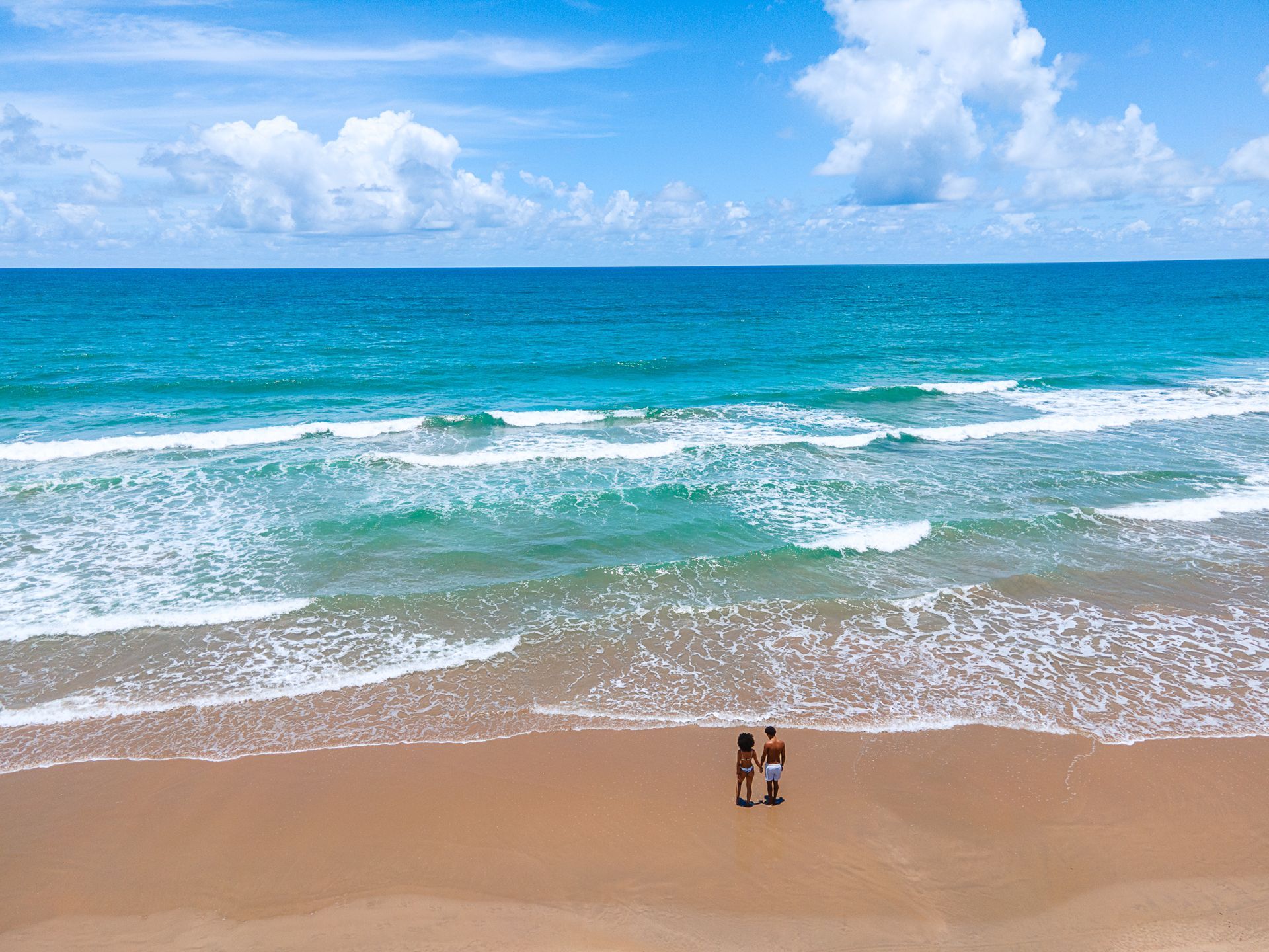 Vista aérea de um casal caminhando à beira-mar em uma praia de águas azuis e céu com nuvens claras.