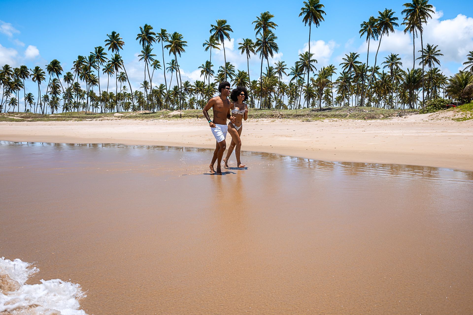 Casal caminha na beira-mar com coqueiral ao fundo, areia molhada refletindo o céu azul em Porto de Galinhas.