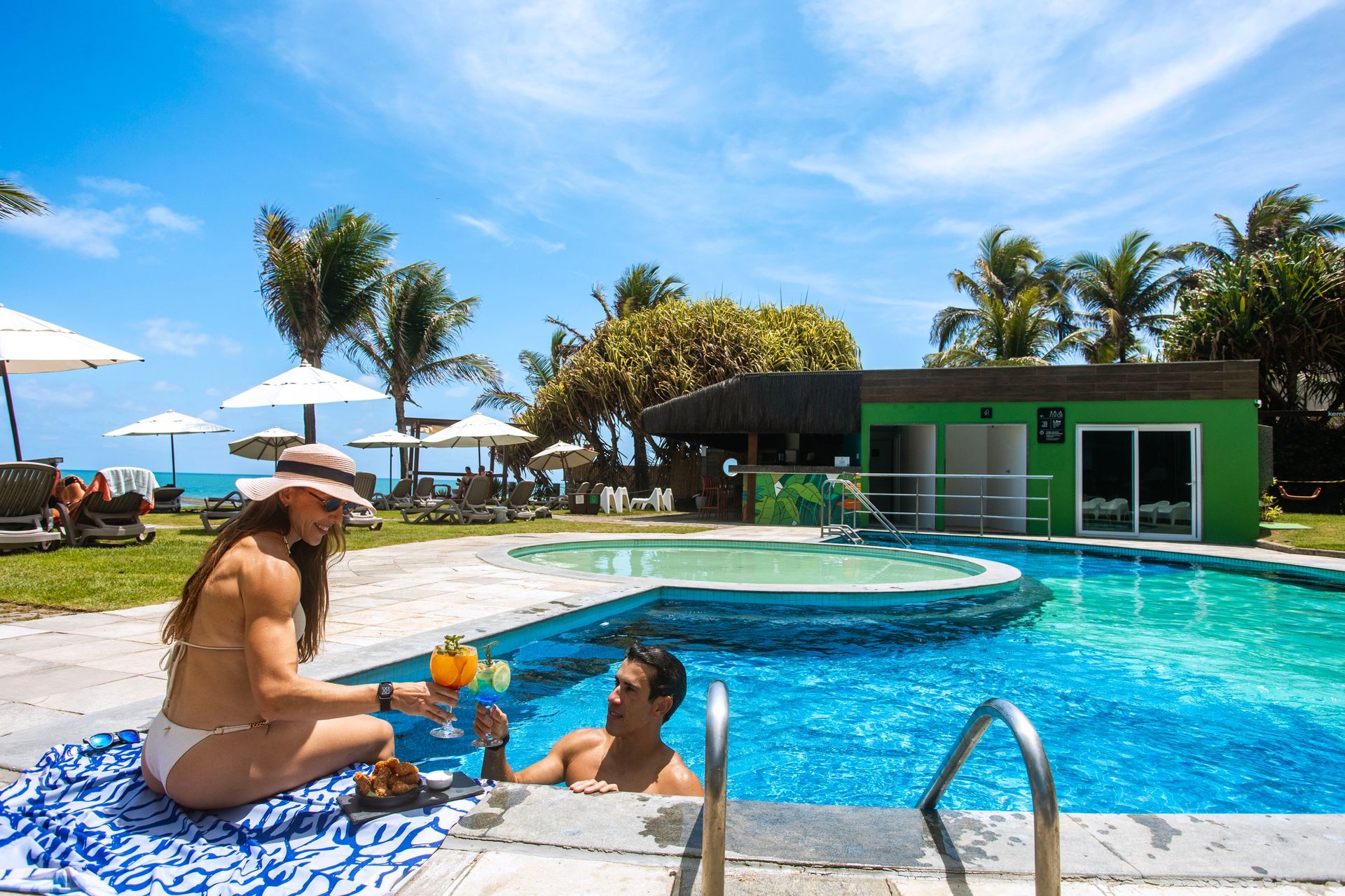 Casal aproveita a piscina à beira-mar com drinks e petiscos em um dia de sol no Kembali Hotel.