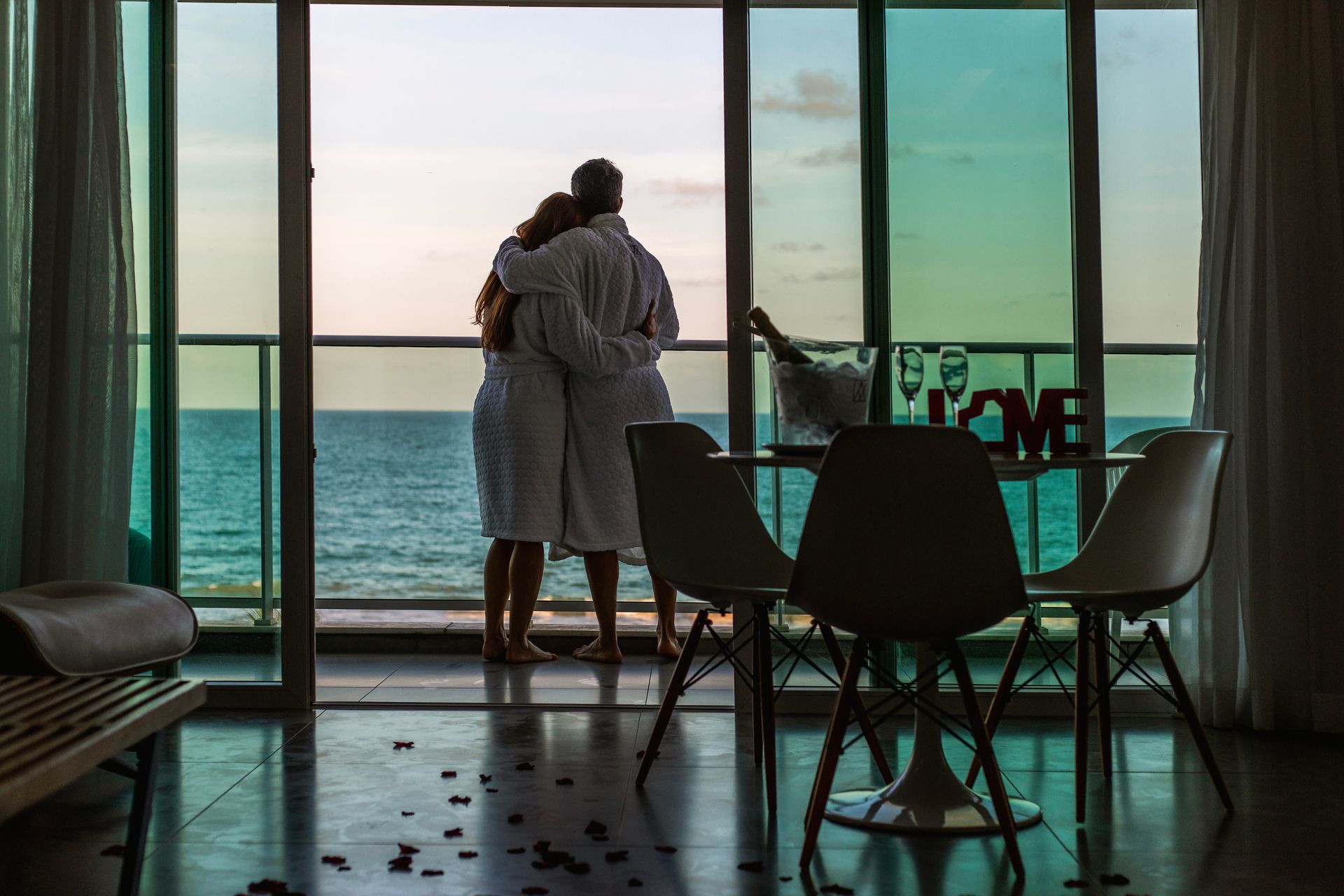 Casal abraçado diante da varanda com vista para o mar, em clima romântico e acolhedor.