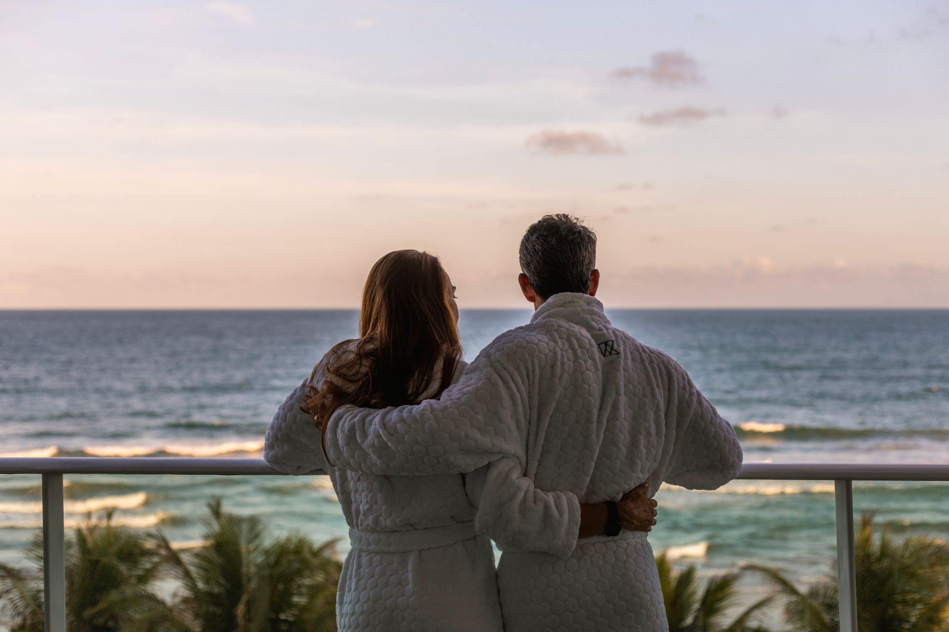 Casal contempla o mar ao entardecer, abraçado na varanda e vestindo roupões.