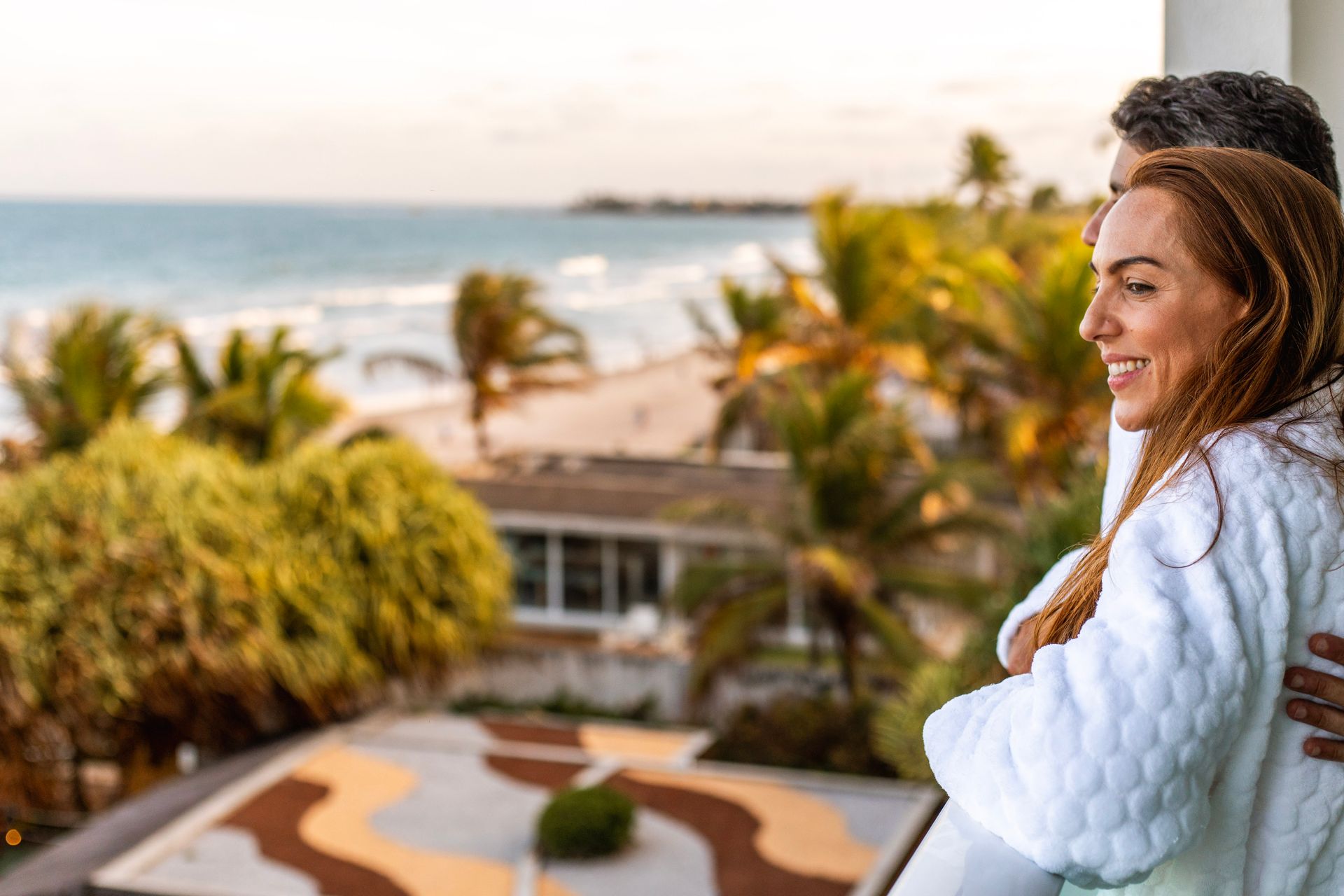Casal de roupão branco sorrindo na varanda com vista para o mar.