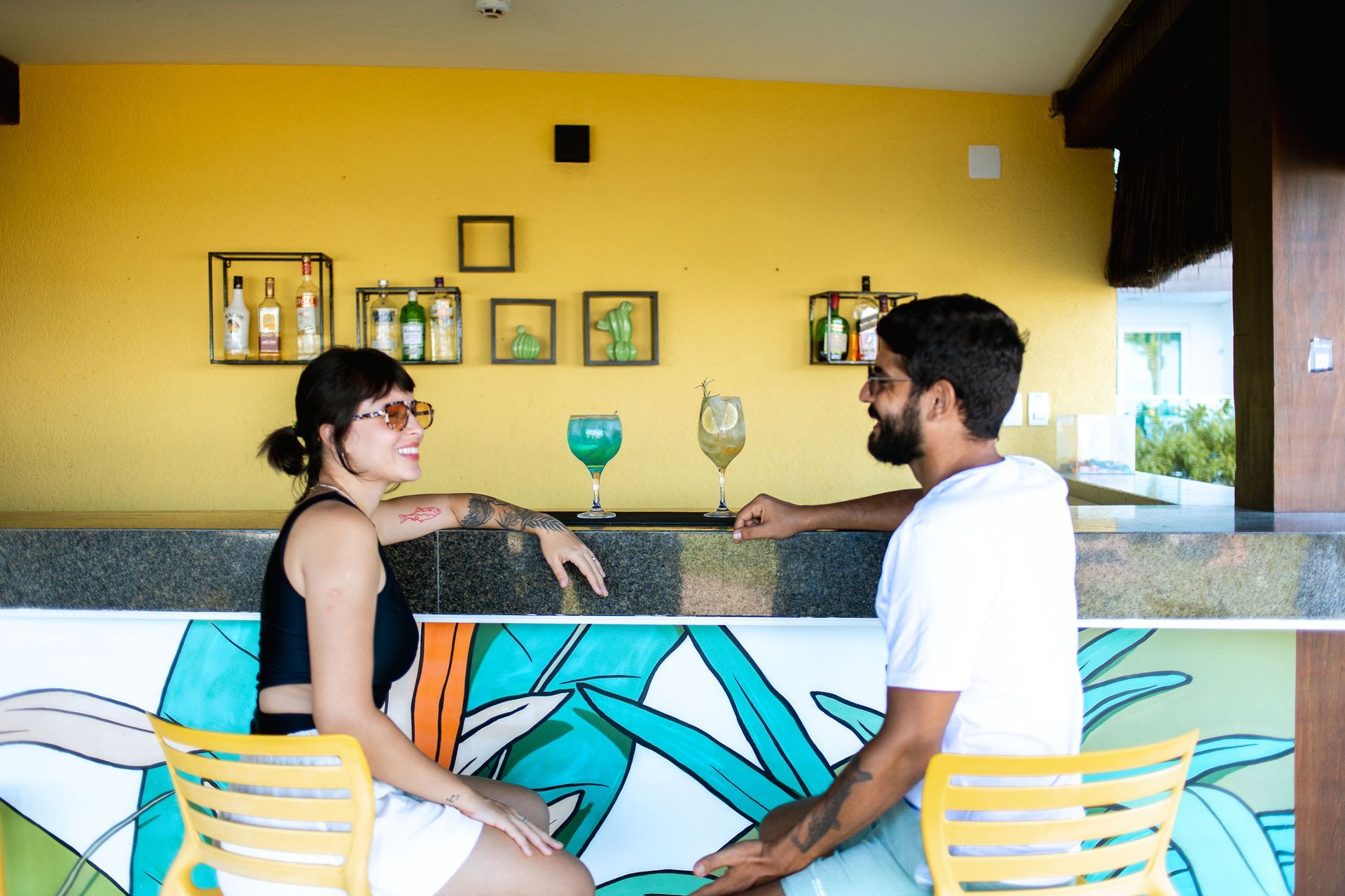 Bar com parede amarela, garrafas em nichos, casal em cadeiras geométricas com drinks coloridos
