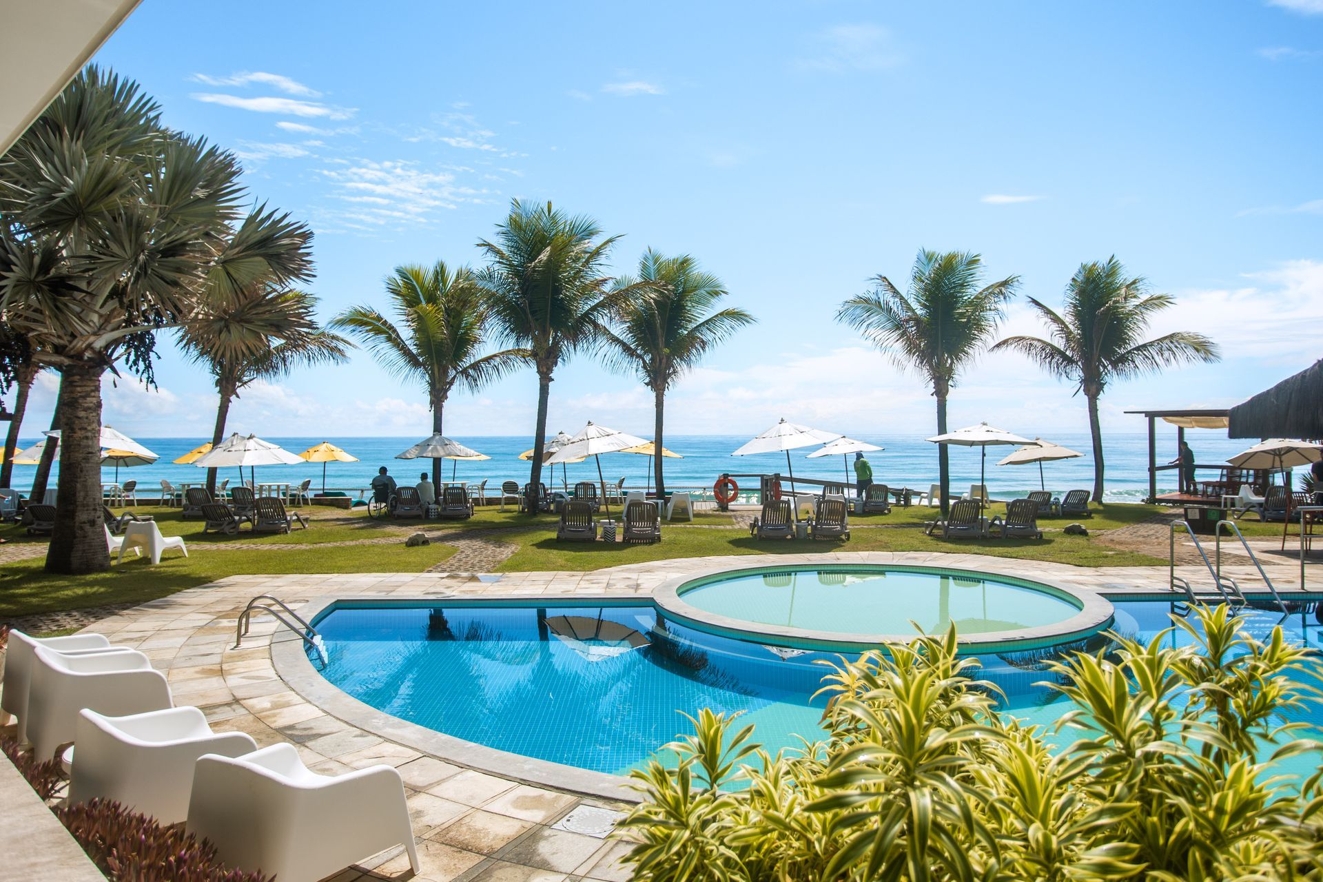 Vista ampla do Kembali, piscina, cadeiras brancas modernas, coqueiros altos, gramado bem cuidado e praia com guarda-sóis brancos & amarelos. Oceano azul & céu claro ao fundo.