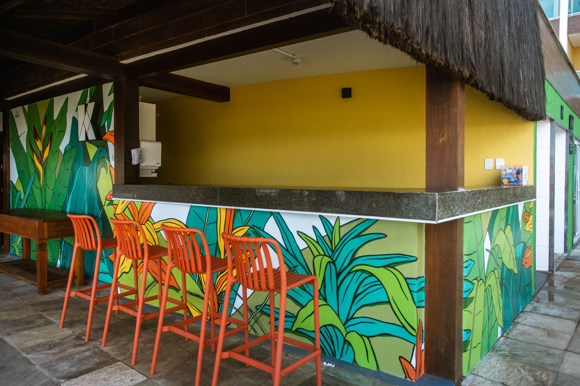 Bar da piscina do hotel com balcão tropical decorado, banquetas coloridas e mural de folhagens em ambiente acolhedor.