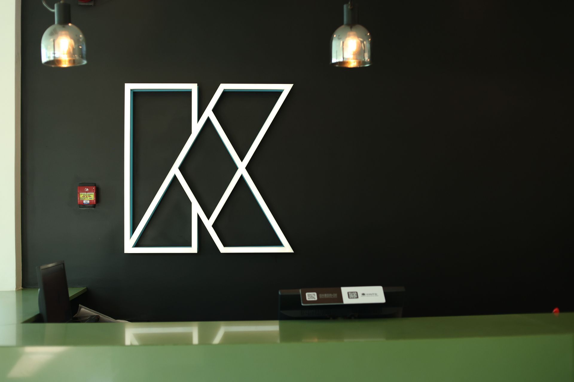 Recepção Kembali com parede verde escuro, logotipo geométrico em neon branco & luminárias pendentes.