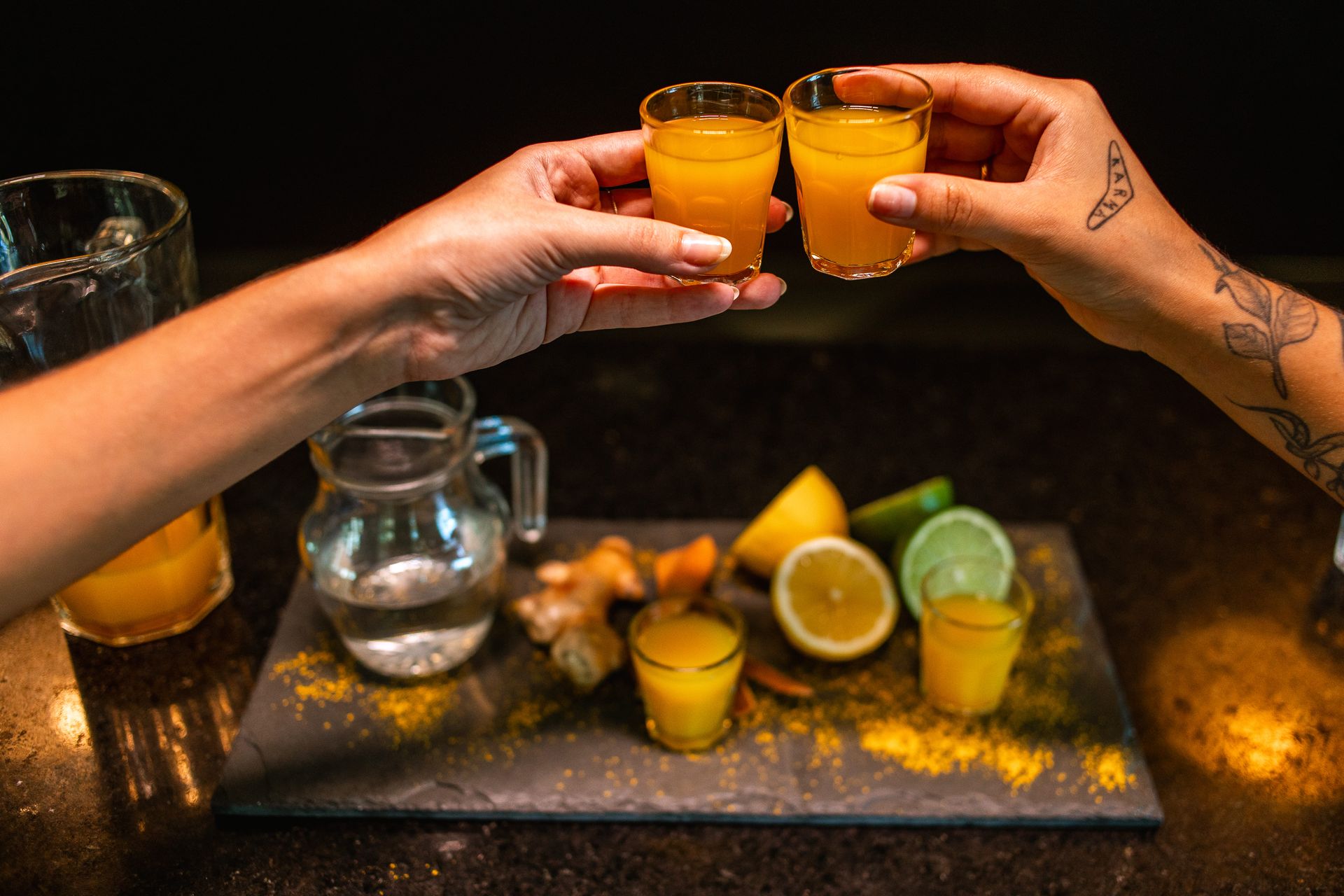 Duas mãos femininas brindando com dois copinhos de vidro de shot amarelo. Ao fundo estão os ingredientes (limão, gengibre) e uma jarra de água.