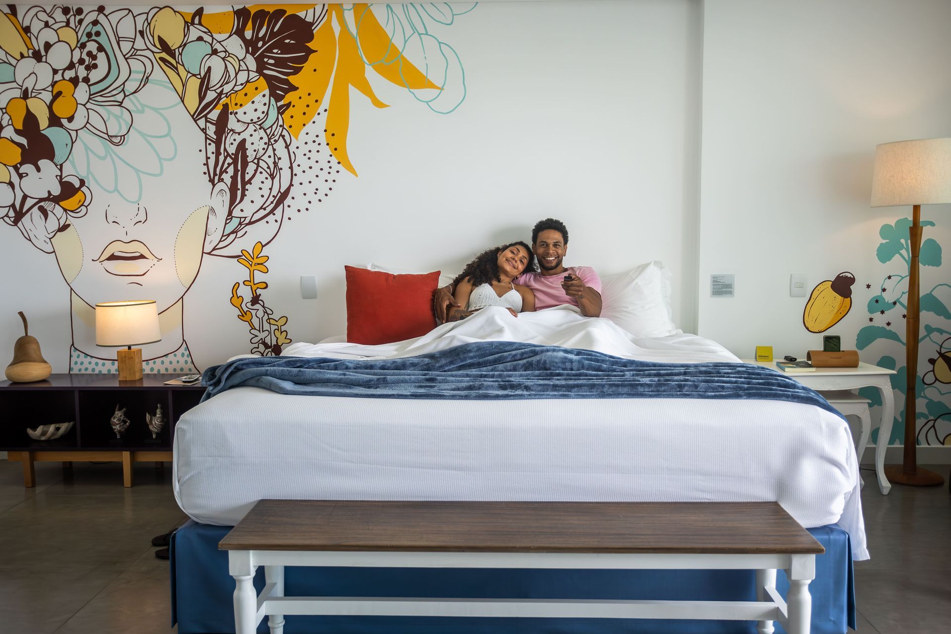 Casal descansando em cama no quarto super do Kembali Hotel. A parede atrás da cama tem uma grande pintura do rosto de mulher, com flores na cabeça. Toda a decoração é bem artística e diferenciada.