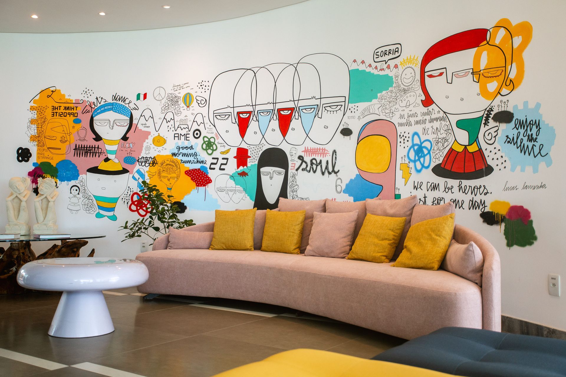 Lobby do hotel, com sofá em tom de rosa claro com várias almofadas em amarelo e rosa claro. A parede atrás possui uma grande pintura colorida em estilo moderno.