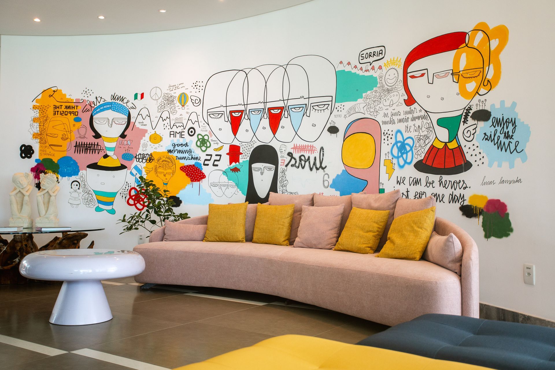 Sofá curvo rosa com almofadas amarelas contrasta com um vibrante e grande mural de arte urbana na parede.
O mural é repleto de desenhos de rostos, figuras contornadas e textos. A decoração é moderna e ousada, focada na expressão artística e cores fortes.