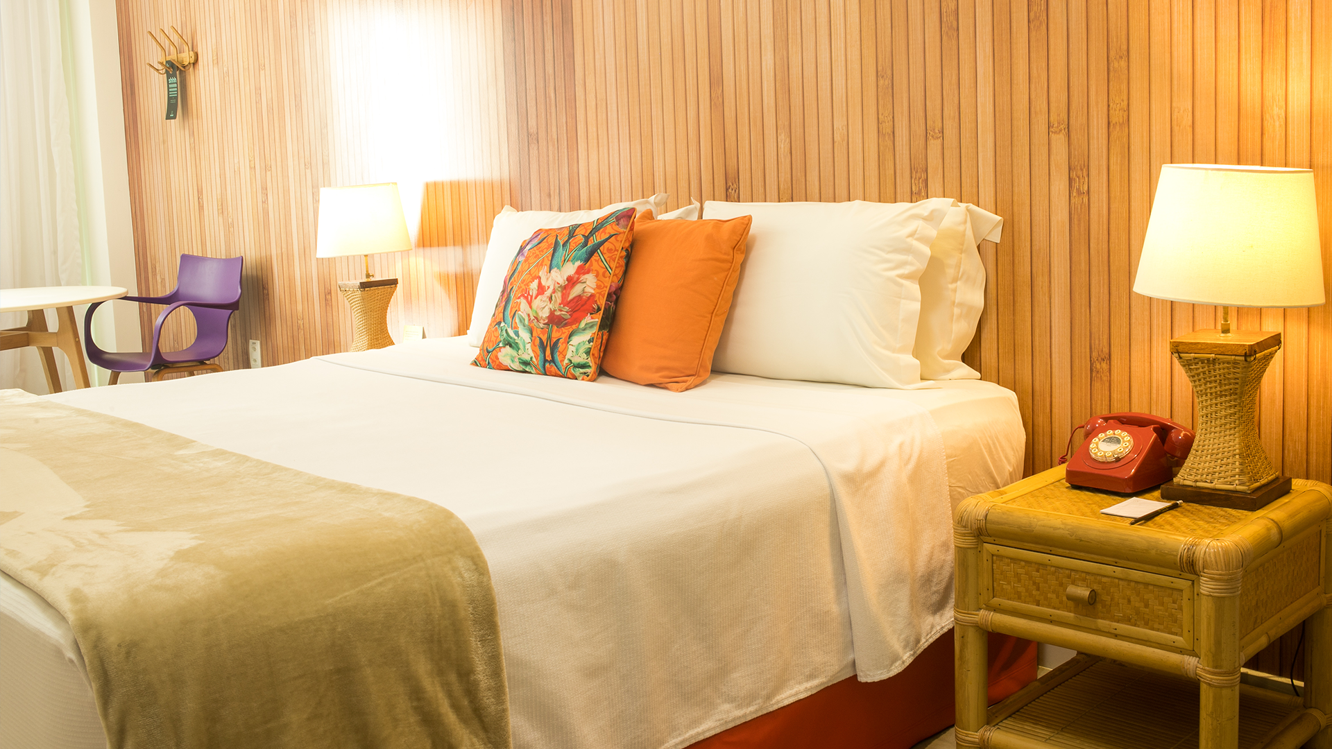 Quarto em estilo praiano, com papel de parede que lembra madeira, cama de casa confortável e móveis em madeira e palha.