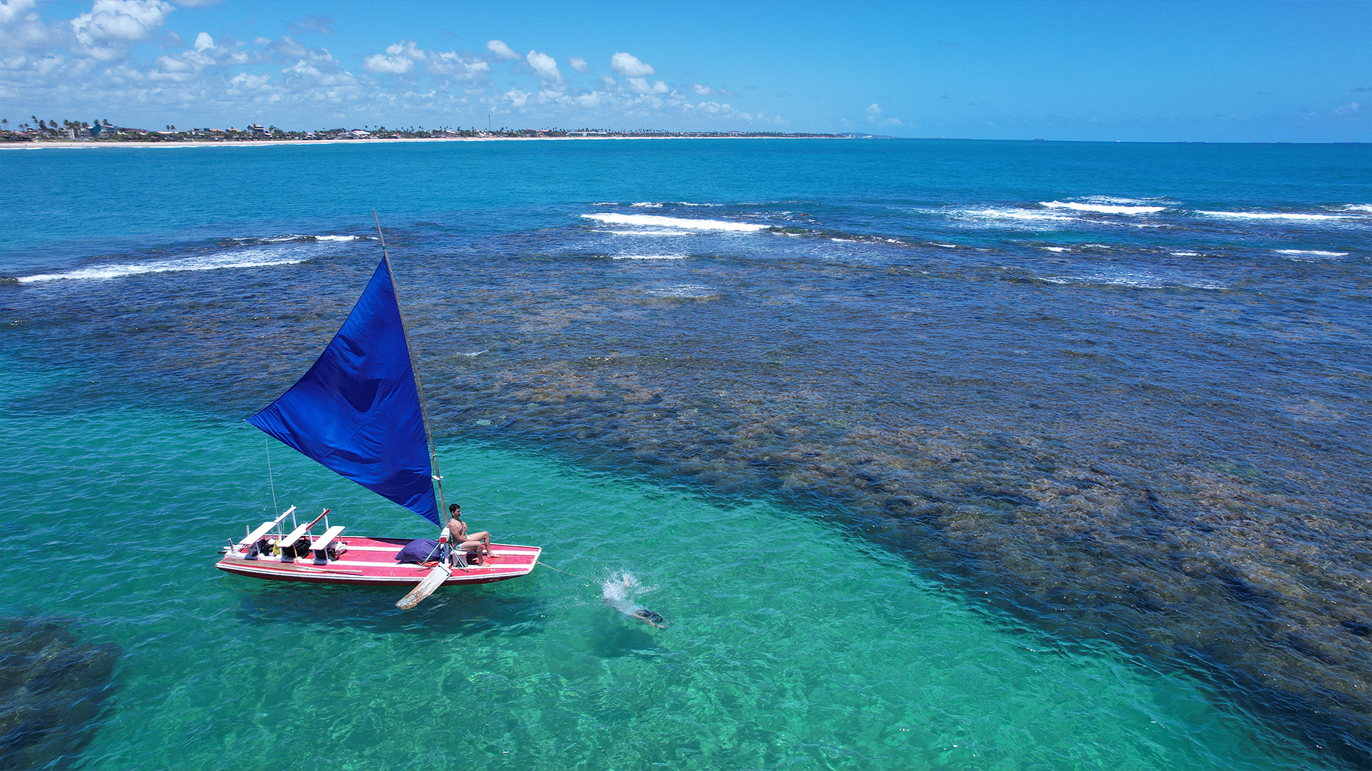 Imagem aérea de uma jangada com vela azul navegando em águas cristalinas sobre os arrecifes em Porto de Galinhas, Pernambuco. A jangada transporta turistas, oferecendo uma vista deslumbrante dos corais submersos em meio ao mar esverdeado.
