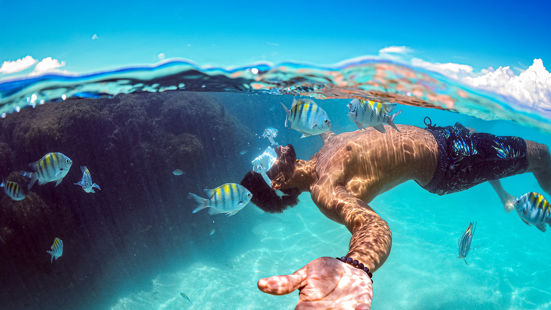 Homem mergulhando nas piscinas naturais, cercado por pequenos peixes. A água é cristalina e os raios de sol batem dentro d'água.