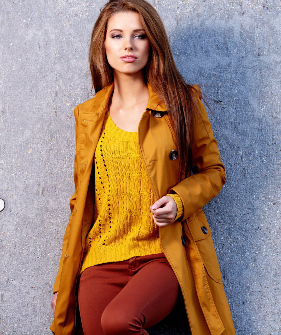 A person wearing a mustard yellow coat and matching knit sweater with dark red pants, standing against a gray wall.
