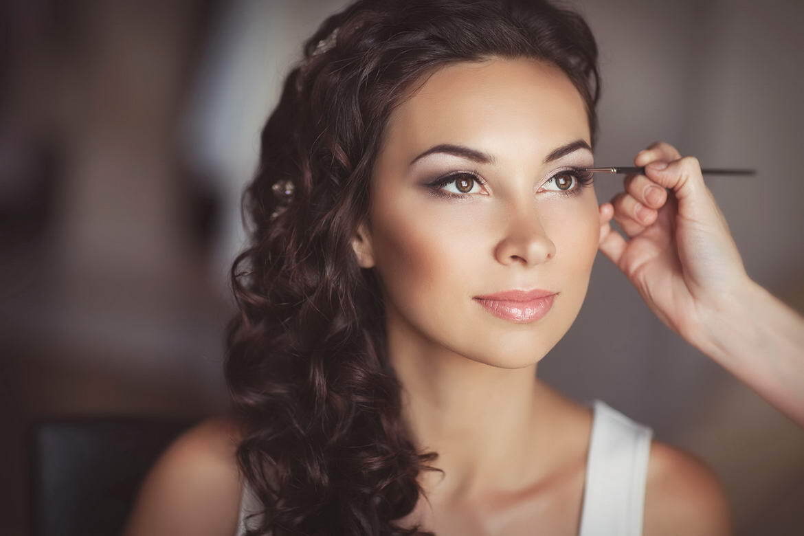 A makeup artist applies eyeliner to a woman with curly brown hair who is wearing a white top.