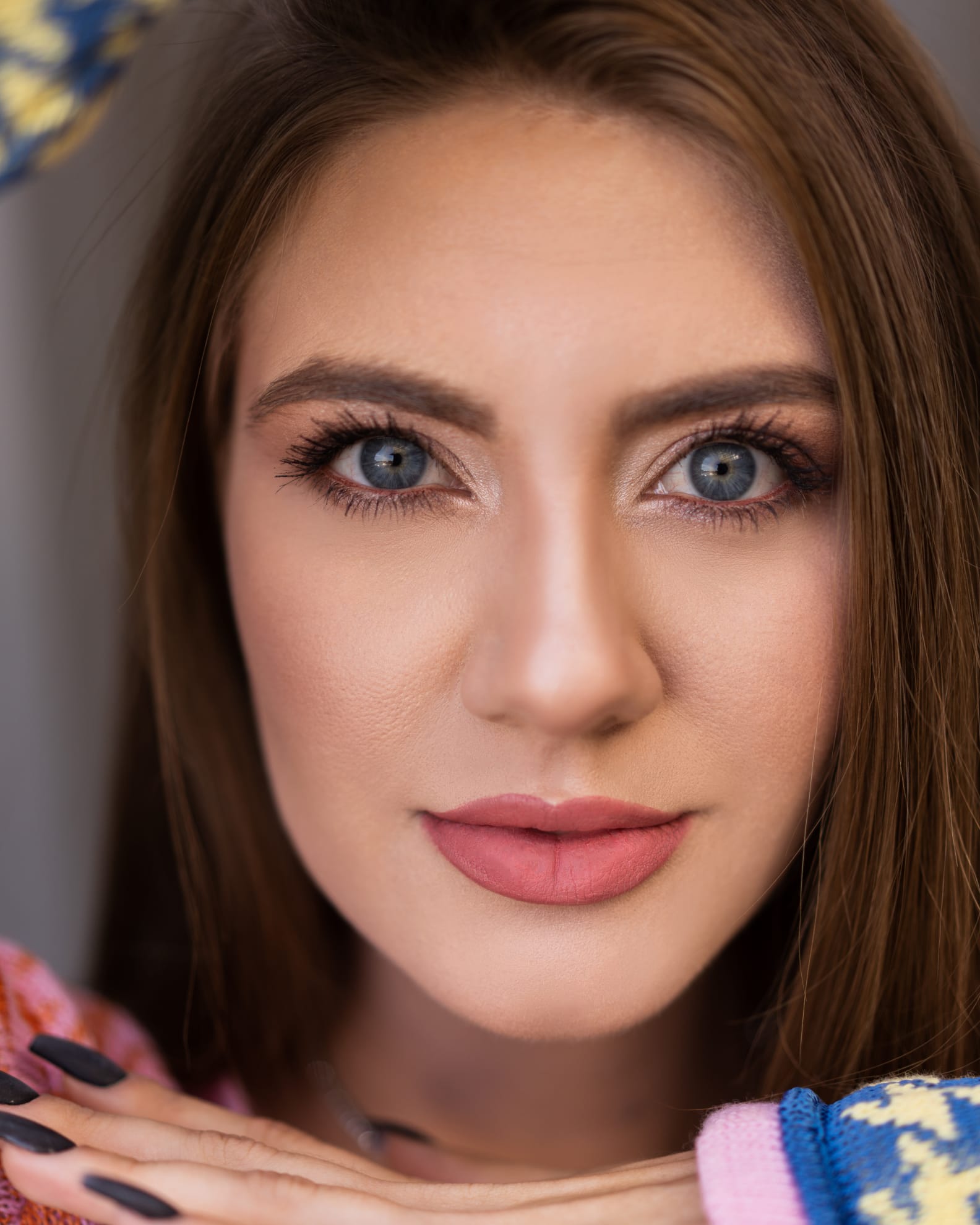 A close-up portrait of a person with blue eyes and light brown hair, wearing a colorful sweater and dark nail polish.
