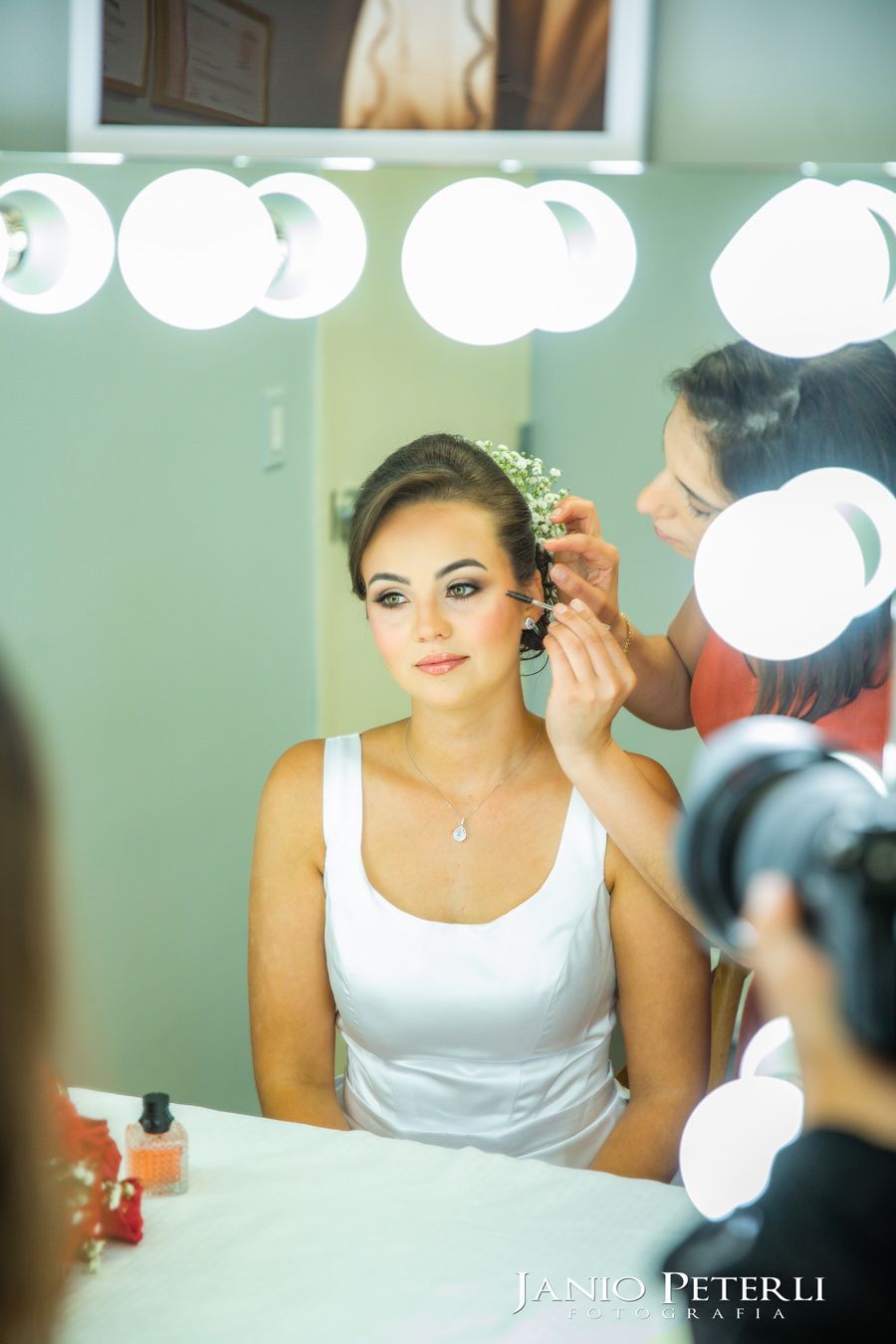 A bride in a white dress sits before a vanity mirror as a hairstylist places a floral accessory into her hair.