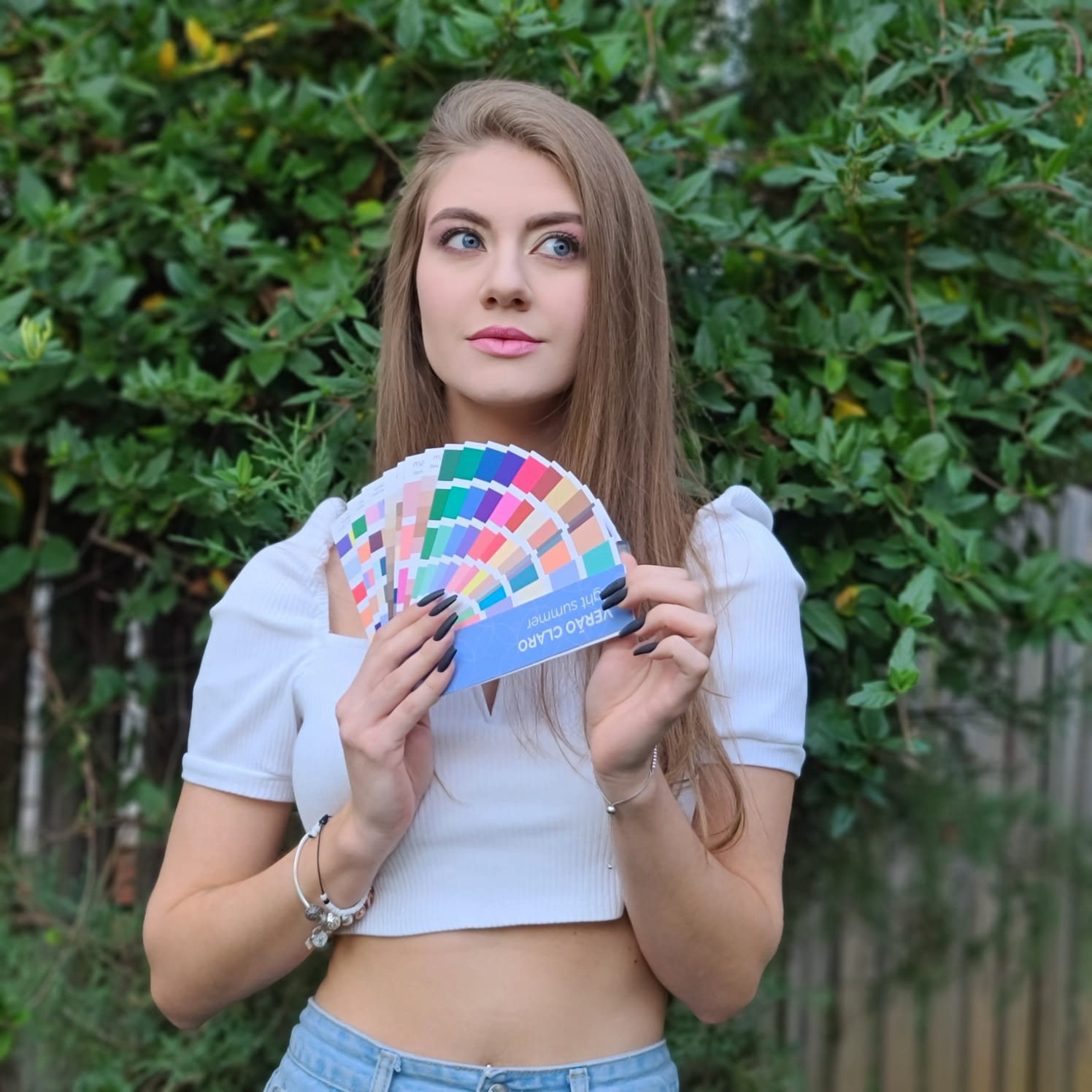 A person holds up a colorful fan of paint swatches in front of a leafy green bush.