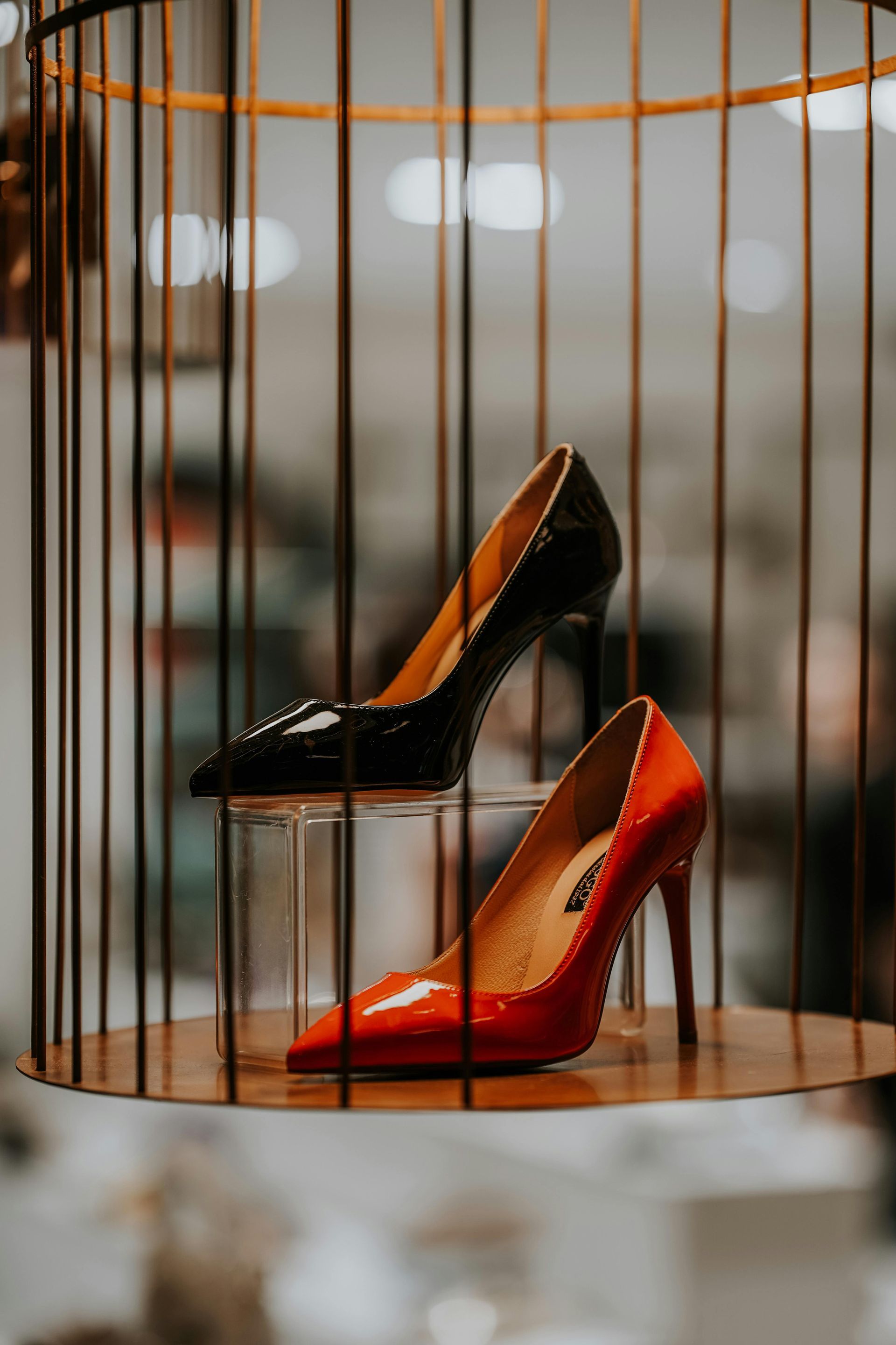 A black high-heeled shoe sits atop a red one, both displayed inside a vertical wire cage structure.