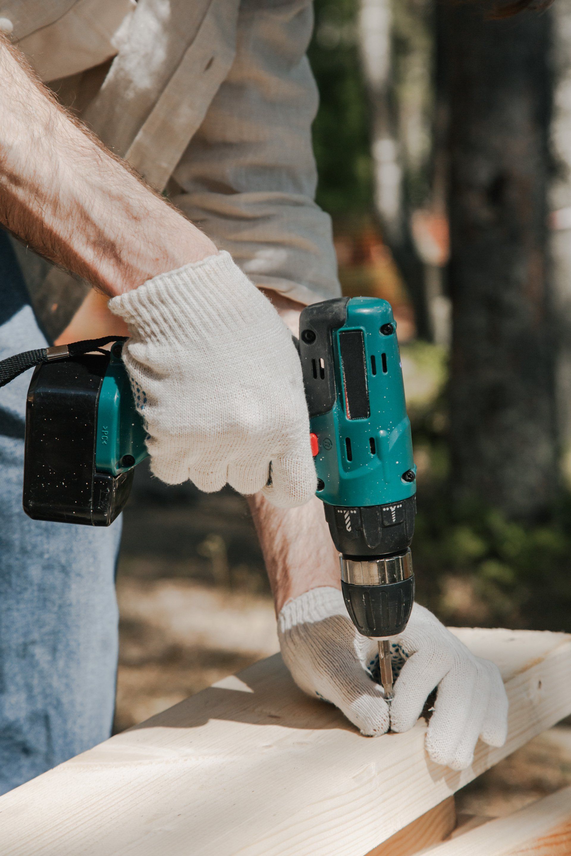 Person using a teal and black drill to screw into a wooden beam, wearing work gloves.