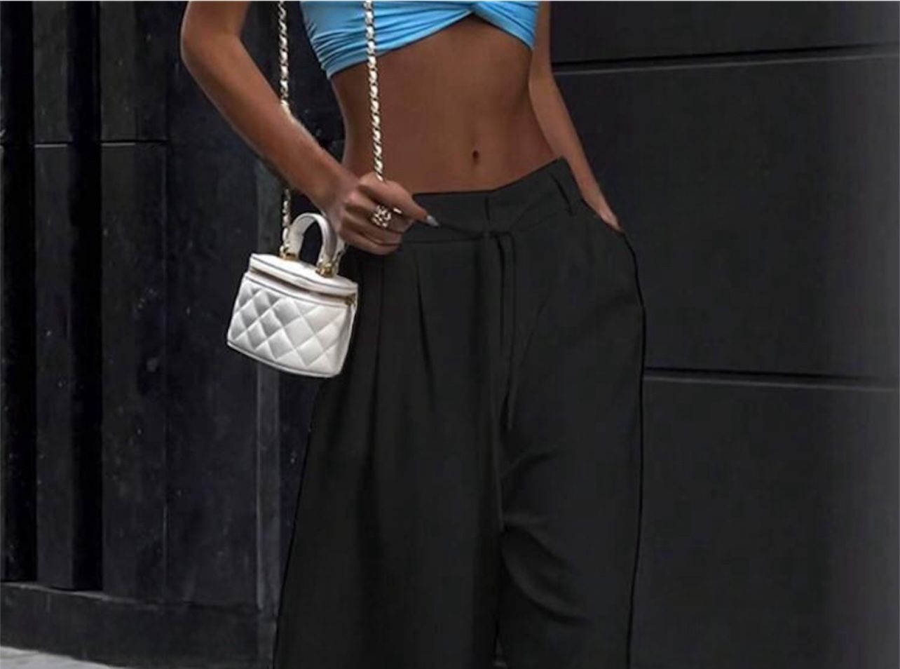 Mujer vestida con un top azul y pantalones negros, sosteniendo un bolso blanco.