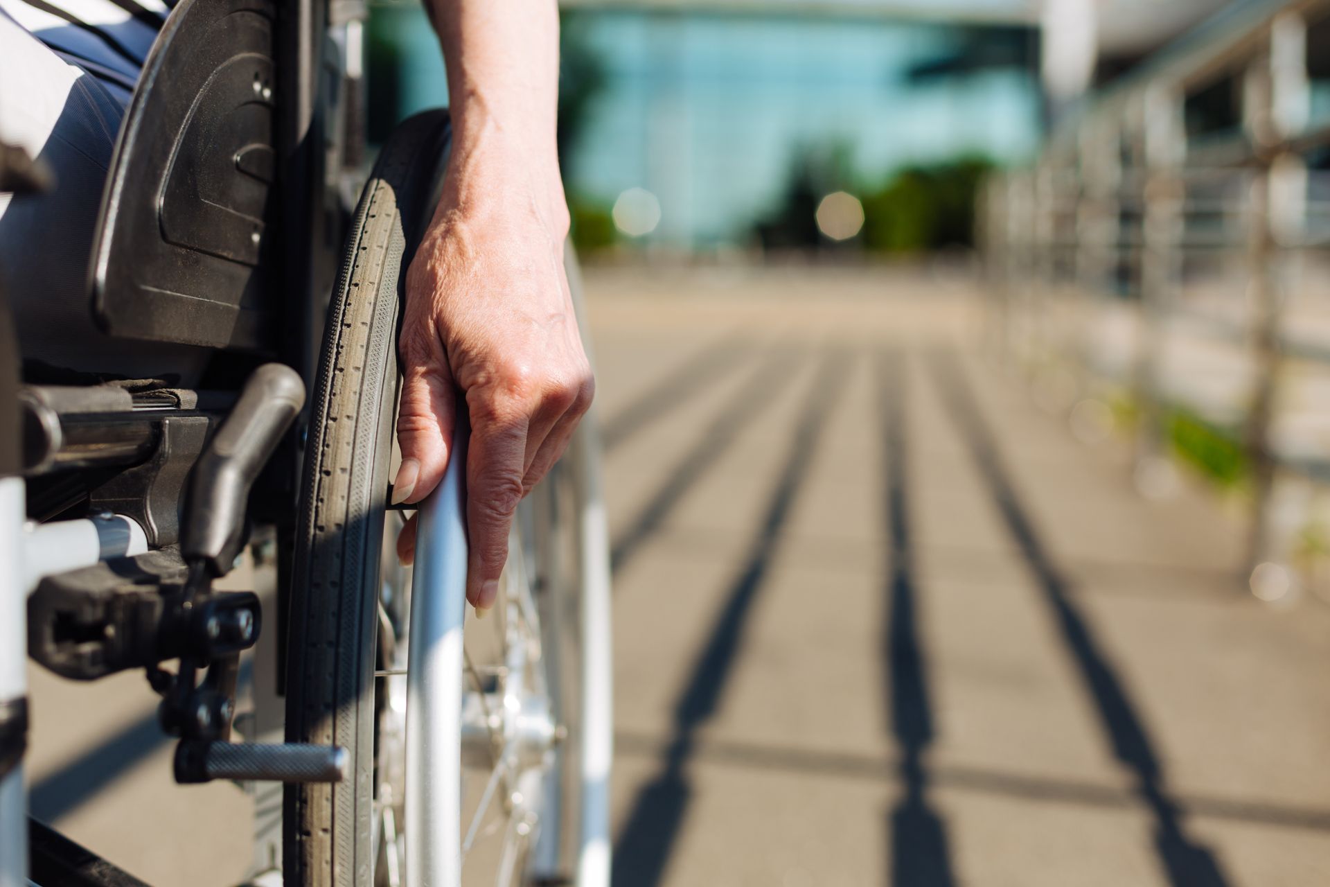 Hand on wheelchair wheel, rolling on accessible ramp, bright sunlight.