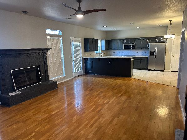 A living room with hardwood floors , a fireplace , a ceiling fan and a kitchen.