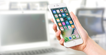 Hand holding a white smartphone with app icons, in front of a blurred laptop workspace.