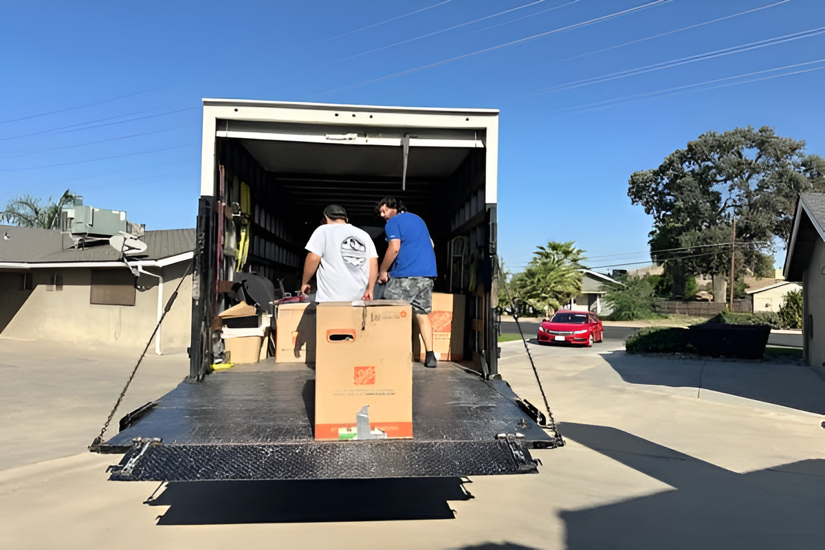 Two people loading boxes into a moving truck, parked on a driveway.