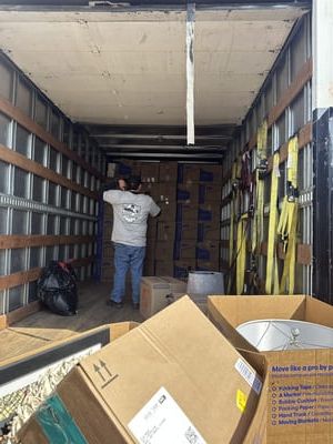 Man in moving truck surrounded by stacked boxes; straps secured to side walls.