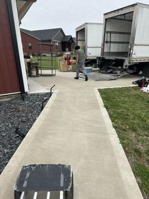 Man walking toward moving trucks on a concrete walkway, boxes and grass on either side.
