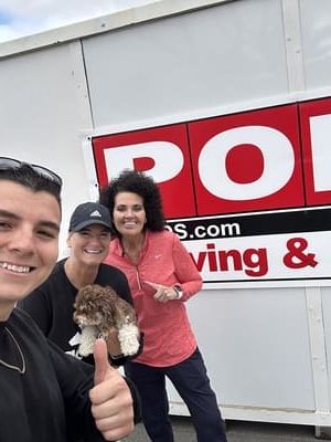Three people smiling by a red and white PODS sign with a dog. One person gives a thumbs up.