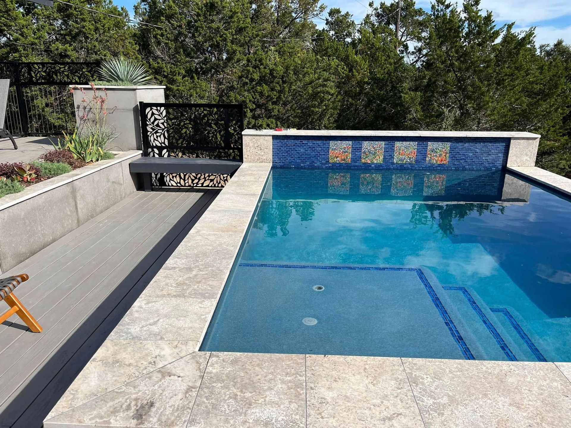 Modern backyard pool with blue water, white stone deck, and decorative tile wall under trees