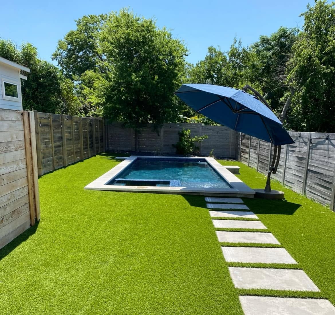 Backyard with artificial grass, small pool, stepping stones, and a blue patio umbrella