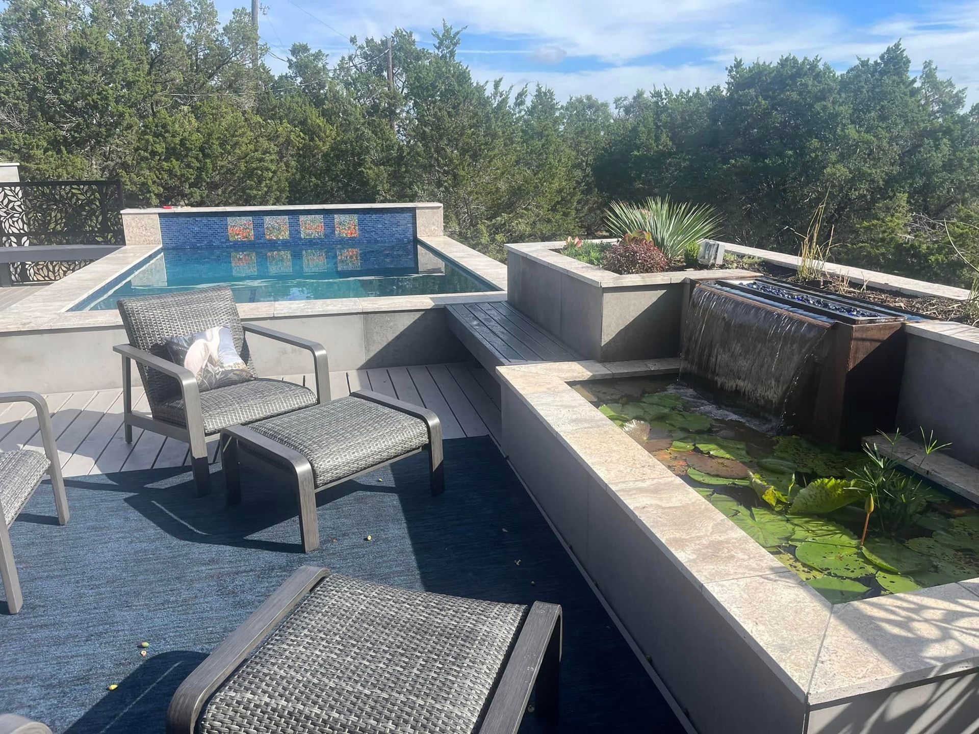 Outdoor patio with chairs, a pool, and a stone planter with greenery under a sunny sky